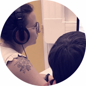 Femme portant un casque audio en pleine session d’enregistrement avec un enfant, devant un micro et un écran d’ordinateur, dans un studio radio chaleureux.